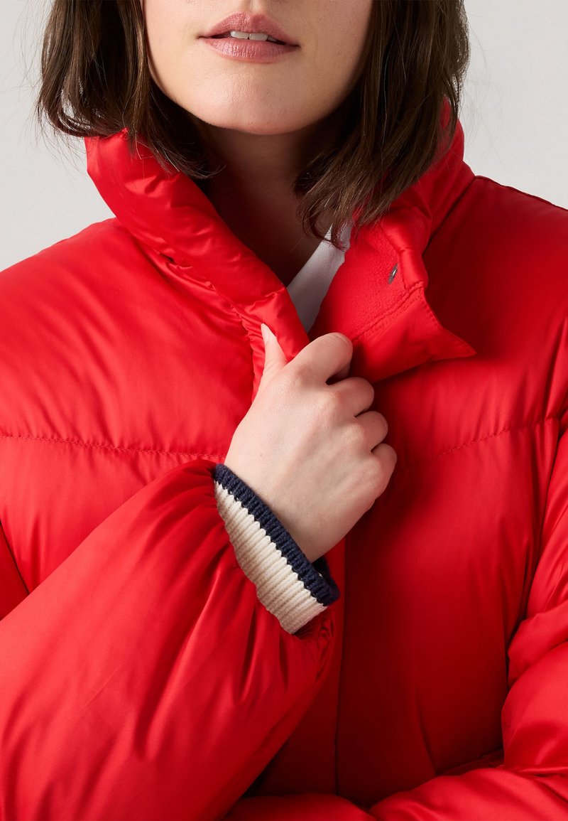 Rote gepolsterte Jacke mit hohem Kragen und gerippten Strickbündchen, die eine glatte Textur und eine enge Passform aufweist und teilweise ein darunter liegendes weißes Hemd verdeckt.