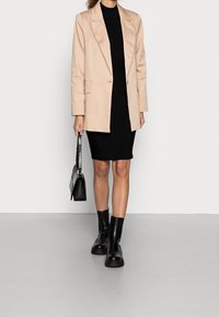 Beige blazer přes černé padnoucí šaty, doplněný černými kotníkovými botami. Blazer má klopový límec a dvě přední kapsy.