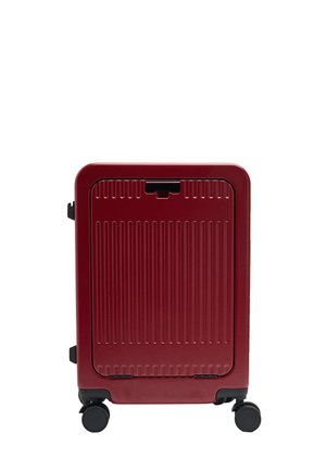 Kompaktowa czerwona walizka z pionowymi rowkami, czterema czarnymi kółkami i przednim panelem dostępowym, stojąca pionowo na białym tle.