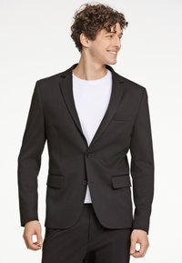 Schwarzer Blazer mit Reverskragen, einem Ein-Knopf-Verschluss und zwei frontalen Taschen. Getragen über einem weißen T-Shirt, das einen taillierten Schnitt hat.
