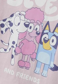 T-shirt graphique en cartoon présentant trois personnages colorés : un chien à taches noir et blanc, un caniche rose, et un chien bleu avec des accents jaunes.