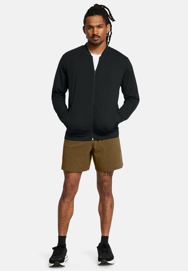 Vêtements d'extérieur noirs zippés avec col montant, associés à un short marron clair. Modèle portant des chaussures de sport noires. Fond uni.