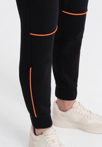 Sorte sweatpants med orange kontrastsyninger, ribbet tekstur på lommerne, tætsiddende manchetter, sammen med lysfarvede sneakers.