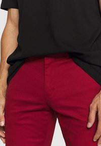 Pantaloni in cotone rossi con vestibilità slim, caratterizzati da gamba dritta, tasche anteriori e una superficie liscia. Abbinati a una camicia nera a maniche corte.