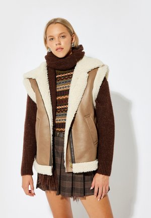 Tan lædervest med hvid pelskrave, sidelommer og lynlåslukning. Brun ribbet turtleneck under, mønstret strik og ternet nederdel.