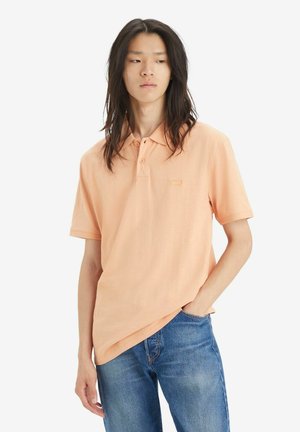 Polo shirt melocotón con cuello, mangas cortas y placket con dos botones; combinado con jeans azules; presenta un sutil logo en el pecho.