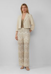Beige cropped jack over een tan top, gecombineerd met wijduitlopende sequined broeken in een chevronpatroon van room- en goudtinten. Lichtbruine sandalen.