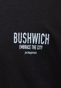 Czarny materiał z białym napisem "BUSHWICH" dużymi, popękanymi literami, poniżej mniejszymi literami "EMBRACE THE CITY" i na dole "JACK & JONES".