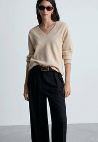 Maglione beige a V in morbido knit con dettagli a coste, abbinato a pantaloni neri a vita alta, fissati con una cintura sottile.