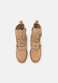 Botas de tornozelo de camurça bege apresentam tiras em camadas, bico arredondado e sola texturizada. Inclui fechos de correr e uma alça traseira para vestir com facilidade.