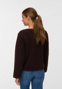 Femme avec une longue queue de cheval brune portant un pull texturé marron foncé et un jean bleu, de dos contre un fond léger uni.