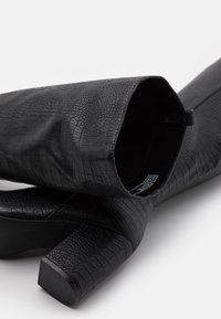 Botas negras hasta la rodilla hechas de materiales texturizados con un patrón de piel de cocodrilo, que cuentan con un tacón grueso y un diseño elegante.