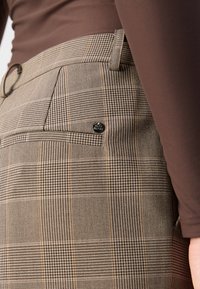 Pantalons à carreaux marron en tissu texturé, avec une poche latérale et un bouton en métal. La taille est étroite avec des passants de ceinture.