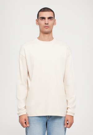 Long-sleeve cream-colored T-shirt, made of soft fabric, featuring a round neck, relaxed fit, and dropped shoulder seams. Worn with light blue jeans.