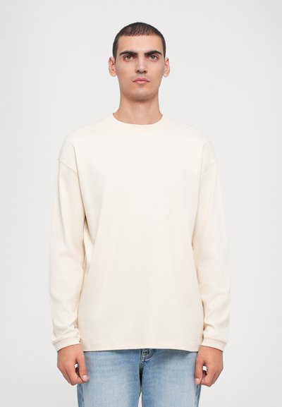 Langärmliges T-Shirt in Cremefarbe, aus weichem Stoff, mit Rundhalsausschnitt, lässiger Passform und überschnittenen Schulternähten. Getragen mit hellblauen Jeans.