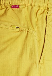 Žluté kraťasy Levi's s texturovanou tkaninou, elastickým pasem, boční kapsou se žlutým zipem a červenou značkovou etiketou.