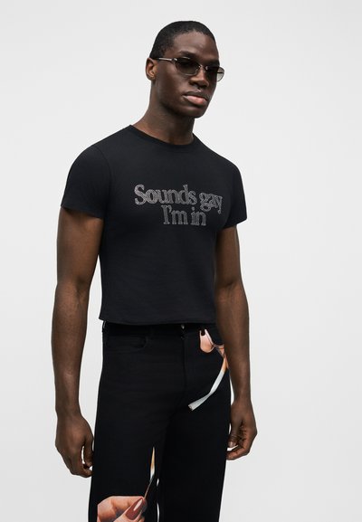Bărbat purtând ochelari colorați, tricou negru scurt cu text din strasuri "Sounds gay I'm in" și pantaloni negri cu mâna imprimată ținând un chibrit.