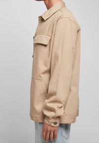 Beige canvasjacka med knappstängning, en bröstficka, sidofickor och manschetter med tryckknappar; avslappnad passform och strukturerad krage.