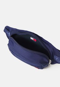 Marsupio in tessuto blu navy con vano principale con zip, tracolla marchiata e una piccola etichetta interna con colori rosso, bianco e blu.