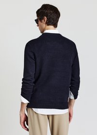 Maglione lavorato a maglia blu navy con scollo rotondo, maniche lunghe e polsini a coste. Indossato sopra una camicia bianca con colletto, presenta una vestibilità comoda.
