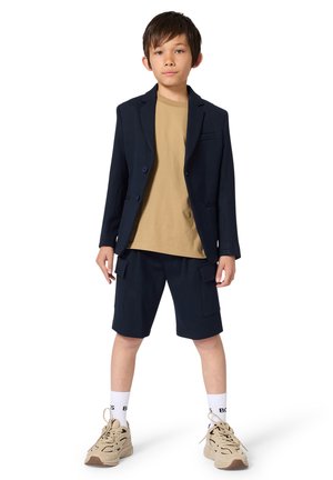 Chłopiec stoi, ubrany w granatową marynarkę, beżowy t-shirt, granatowe krótkie spodnie typu cargo, białe skarpetki i beżowe trampki, na białym tle.