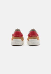 Tenisky s tělem z semiše v tan barvě, červenými akcenty na patě a bílou gumovou podrážkou. Mají ražené logo na zadní straně.