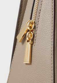 Détails d'un sac en cuir tan avec une fermeture éclair en or. Présente des tirettes de fermeture éclair doubles et une surface texturée, mettant en avant le savoir-faire et la qualité des matériaux.