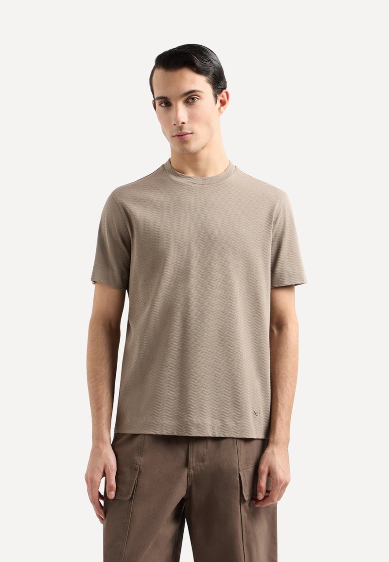 T-shirt a maniche corte, di colore marrone chiaro, con un motivo a onde texturizzato e scollo a girocollo, abbinato a pantaloni cargo marrone scuro.