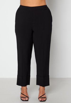 Vrouw draagt zwarte cropped rechte broek met grote omslagen, gecombineerd met zwarte sandaal met banden en hakken op een lichte achtergrond.