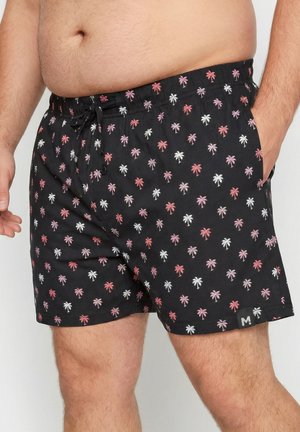 Nærbillede af en person, der bærer sorte badeshorts med et mønster af rosa og hvide palmetræer, hånden i lommen og elastik i taljen med snøre.