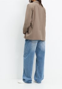 Person wearing a beige blazer, loose light blue jeans, and white sneakers standing against a white background, facing away.