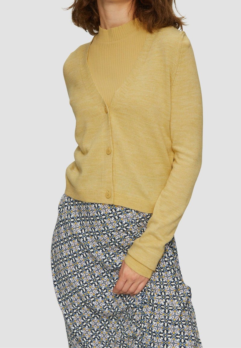 Yellow button-up cardigan over a ribbed mustard top, paired with a patterned skirt featuring a floral design in blue, white, and yellow tones.