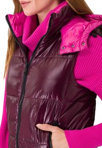 Femme portant un gilet matelassé bordeaux brillant sur un pull à col montant côtelé rose vif, la main dans la poche du gilet.