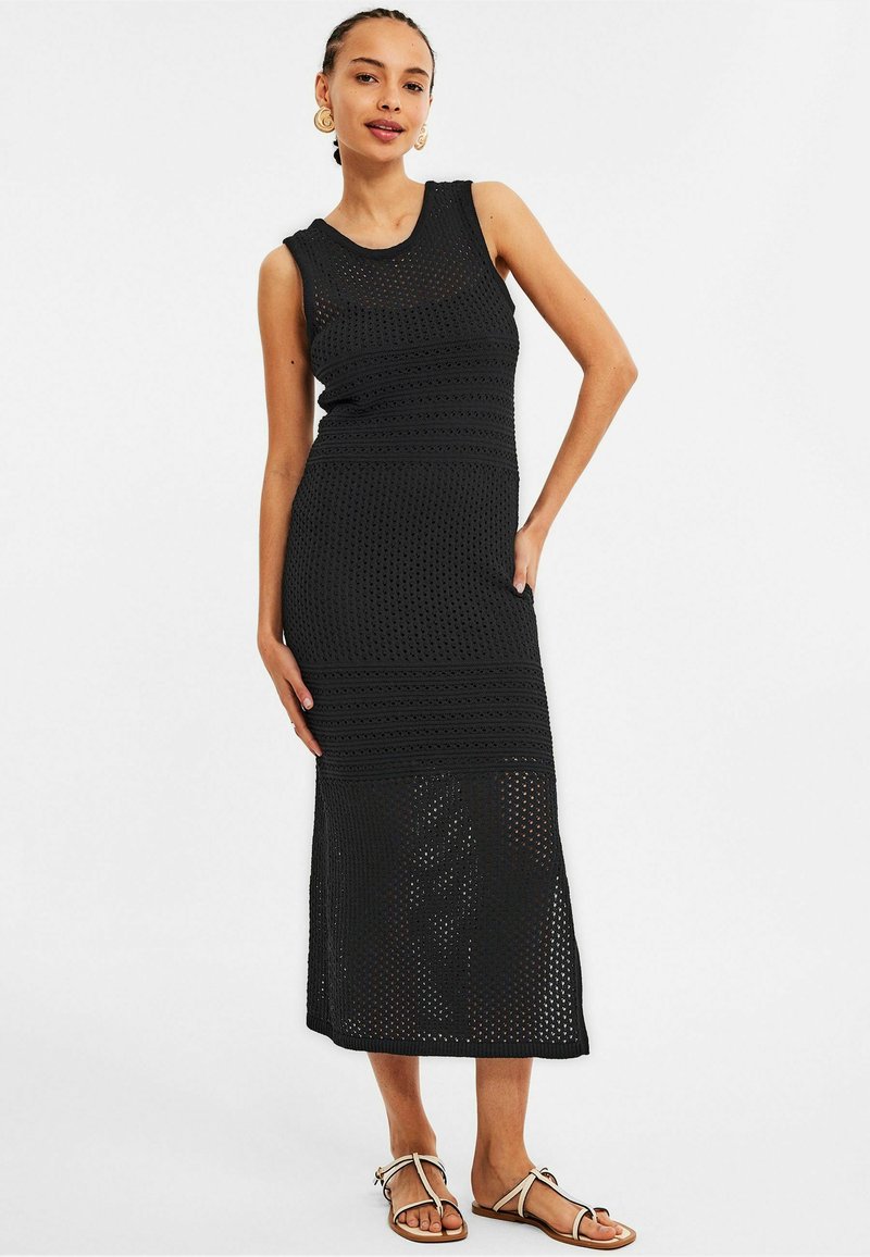 Robe sans manches en tricot noir avec un motif en mailles ouvertes, longueur genou, bandes texturées et silhouette ajustée. Associée à des sandales plates.