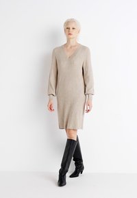 Abito lavorato a maglia beige con scollatura a V, maniche lunghe e un taglio morbido, abbinato a stivali alti neri in pelle. Texture morbida e design minimal.