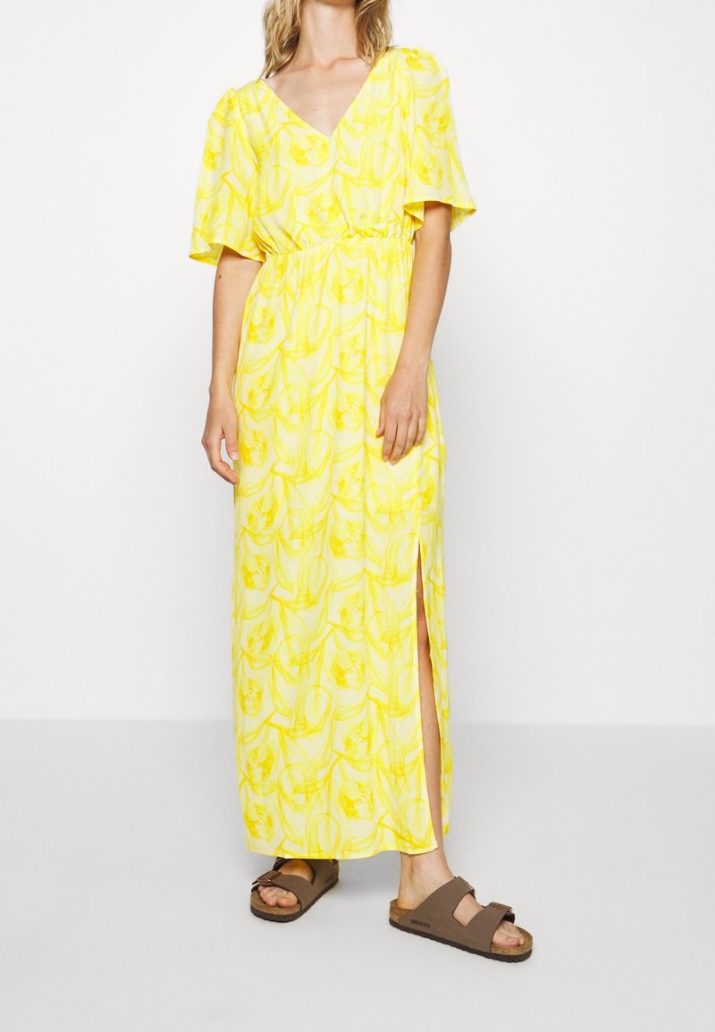 Robe maxi florale jaune avec des manches courtes bouffantes, décolleté en V, taille élastique et fente sur le côté. Associée à des sandales marron.