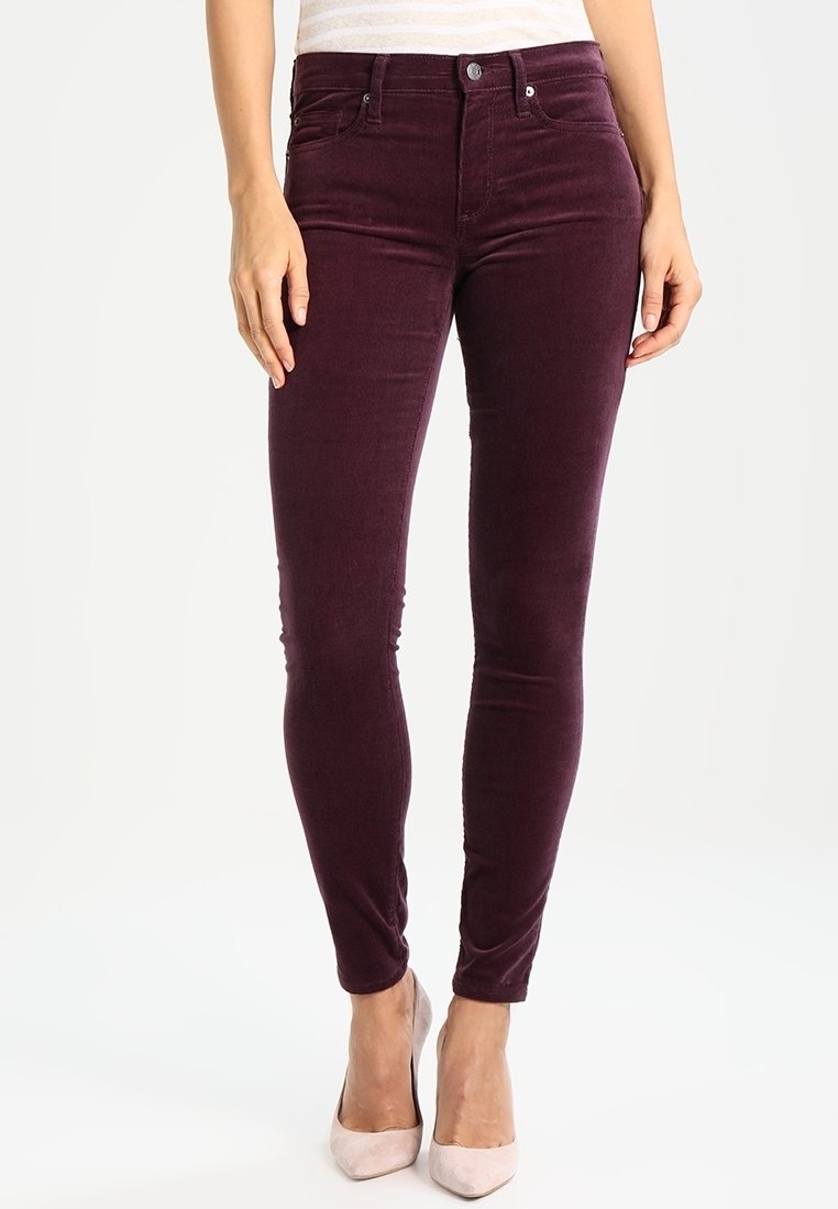 Femme portant un pantalon en velours slim violet foncé et des chaussures à talons hauts beige clair, debout les mains détendues sur les côtés.