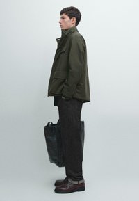 Giacca impermeabile verde oliva con due tasche frontali, indossata sopra pantaloni grigio scuro. Borsa tote nera e scarpe marroni con suole testurizzate.