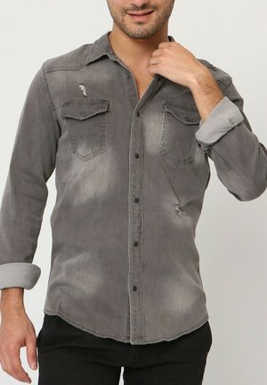Chemise en denim grise avec deux poches poitrine, détails usés, boutons noirs et manches retroussées. Texture douce et conception à ourlet arrondi.
