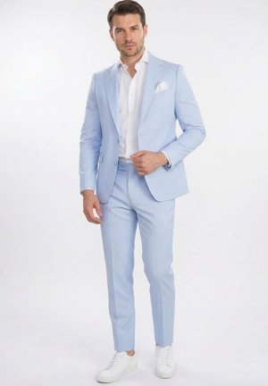 Hombre con traje azul claro, camisa blanca y zapatillas blancas, de pie frente a un fondo blanco.