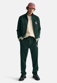 Zielony dres z białymi paskami i logo, zestawiony z kremowym swetrem w warkocze i różową czapką, na jednolitym tle.