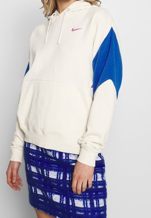 Persoon die een crèmekleurige Nike-hoodie met blauwe mouwen en een blauw-witte geruite rok draagt, staand tegen een effen achtergrond.