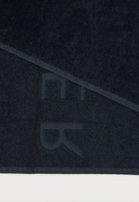 Πετσέτα σε ναυτικό μπλε χρώμα με υφή στην επιφάνεια, που διαθέτει μία γωνία με λεία επιφάνεια και ανάγλυφο λογότυπο. Οι άκρες είναι περιποιημένα ραμμένες.