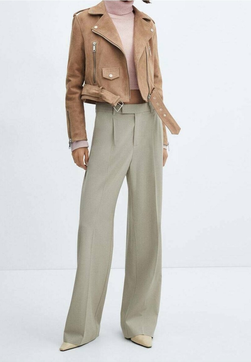 Cropped Motorradjacket aus braunem Wildleder mit silbernen Reißverschlüssen, kombiniert mit olivegrünen, hochgeschnittenen, weit geschnittenen Hosen und spitzen beigen Schuhen.