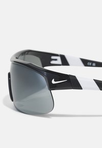 Sports saulesbrilles ar melnu rāmi, pelēkiem lēcu tonējumiem un baltām akcentiem uz kājām, kurām ir Nike logo. Teksturēta tempļa dizains.