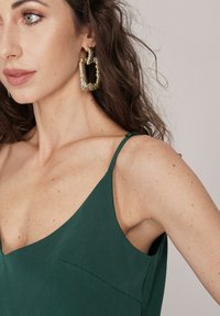 Ciemnozielona, bezrękawnikowa sukienka z dekoltem w kształcie litery V. Wykonana z gładkiego, teksturowanego materiału. Złote, skręcone kolczyki w kształcie kół dodają szczegółu.
