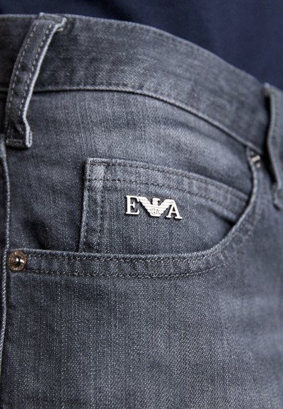 Jeans in denim grigio scuro con tasca frontale dotata di un pin argentato con il logo 'EVA' e dettagli di cucitura visibili sulla cintura e sulle tasche.