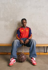 Ung man i spansk fotbollsjersey och röda sneakers sitter på en bänk och håller en vintage läderfotboll i en nätpåse mot en betongvägg.