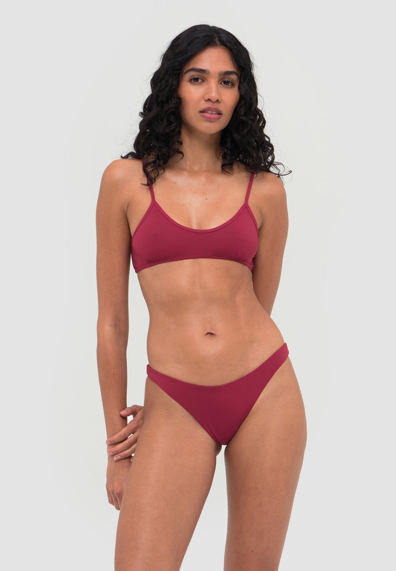 Femme aux cheveux bouclés foncés portant un bikini bordeaux à fines bretelles, debout devant un fond gris clair uni.