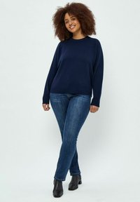 Marineblauer Pullover mit Rundhalsausschnitt, lockerer Passform und langen Ärmeln, kombiniert mit dunkelblauen Slim-Fit-Jeans und schwarzen Knöchelstiefeln.